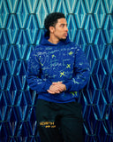 Model wearing the Cobalt Blue Graffiti Hoodie against a blue patterned wall, showing the hand-painted graphic