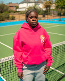Front view of model wearing the North Bioko Club Peach Hoodie in Hibiskus Pink on a tennis court