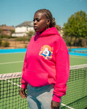 Model wearing the North Bioko Club Peach Hoodie in Hibiskus Pink, three-quarter view on a tennis court